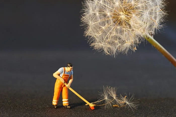 Toy working in high-visibility clothing sweeping up the seeds of a dandelion. The dandelion is looming over the worker.